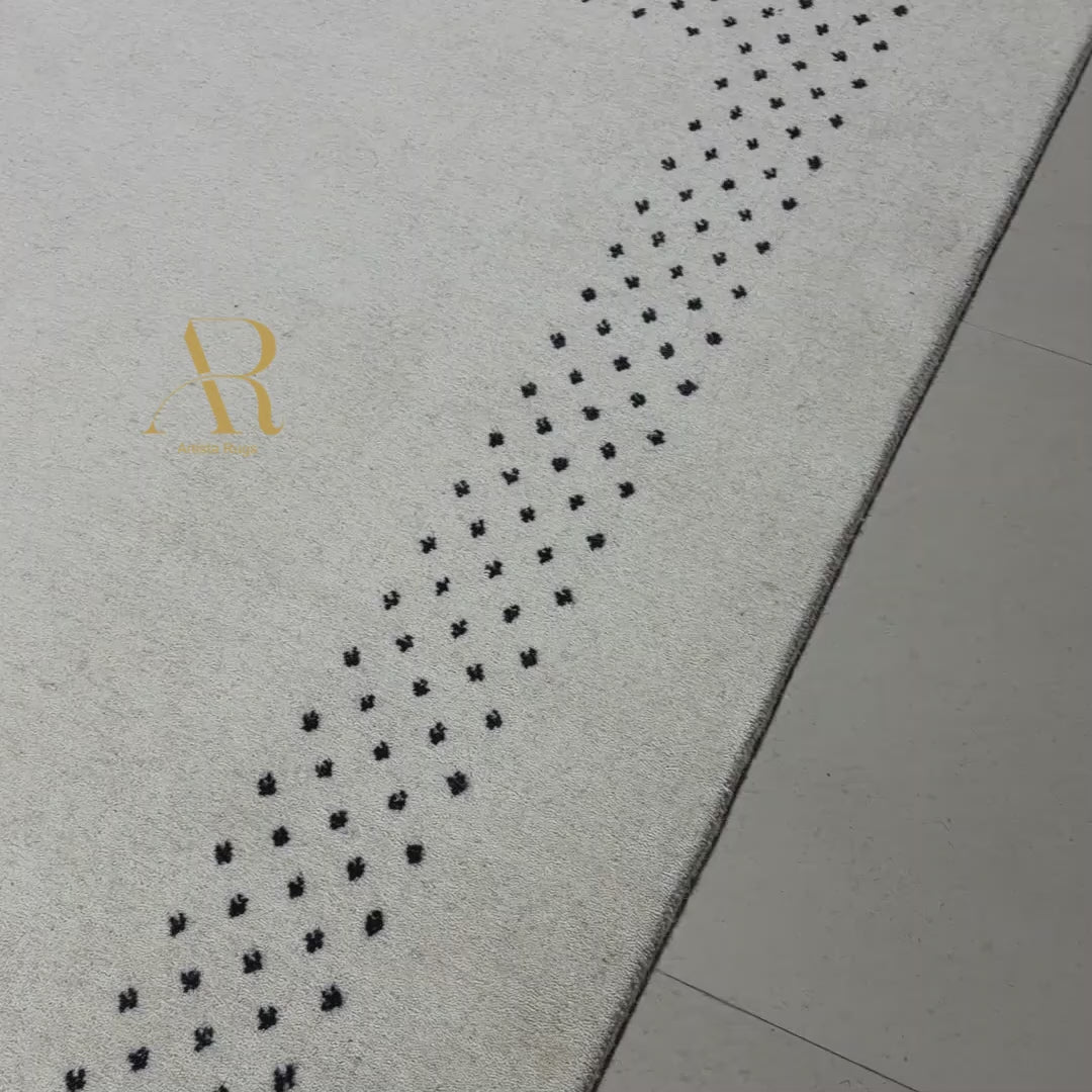 Close-up of a textured light beige rug with a repeating pattern of small black floral or clover-like motifs arranged in a curved row.