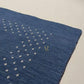 A dark navy blue rectangular rug with a textured surface, featuring a decorative border of small white dots and a gold monogram logo in the center, shown against a plain white background.