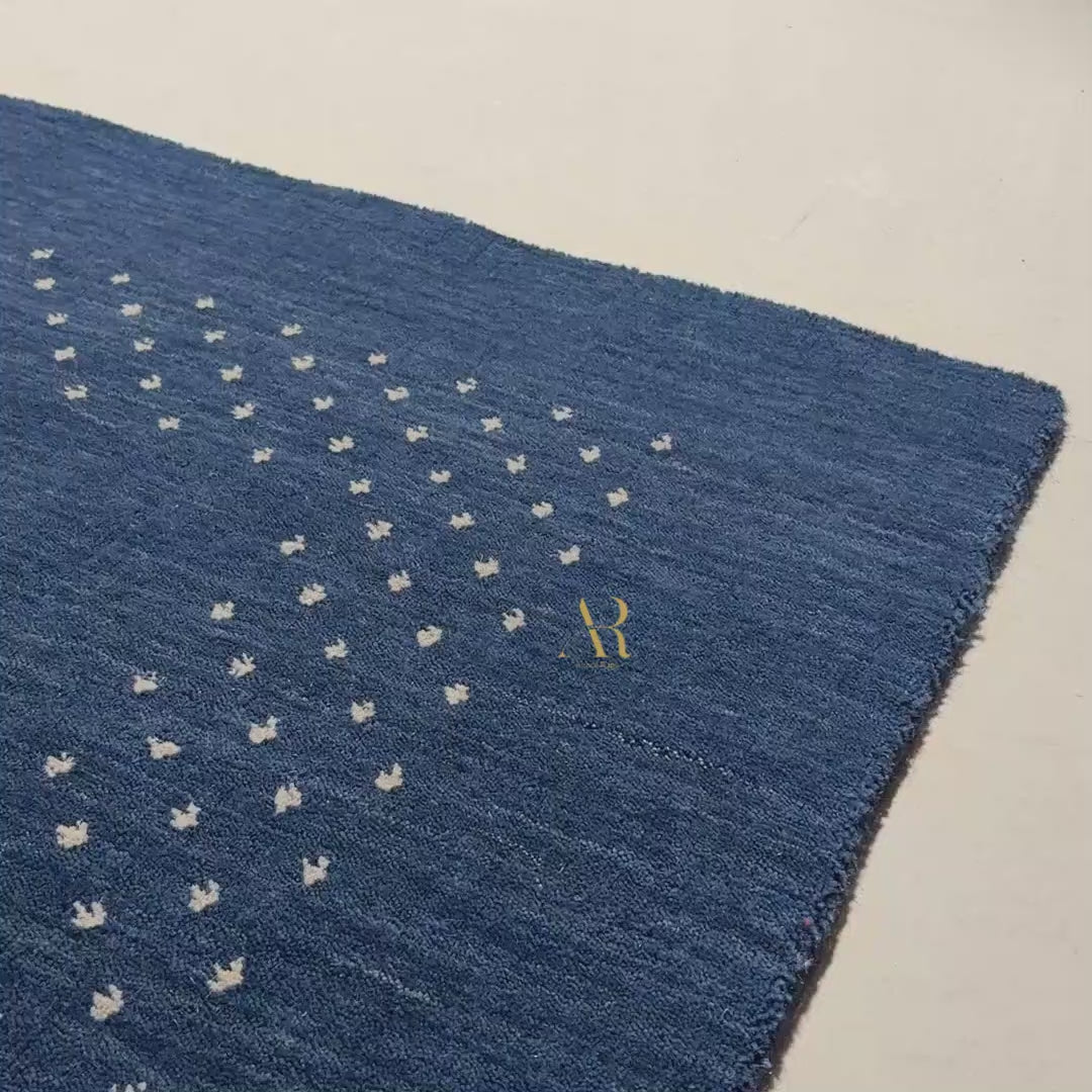 A dark navy blue rectangular rug with a textured surface, featuring a decorative border of small white dots and a gold monogram logo in the center, shown against a plain white background.